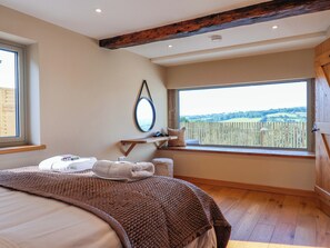 A bedroom with a large bed, a wooden ceiling beam, a window with a view of the countryside, and a bedside table with a mirror and a pillow. - Heathmoor (Yelverton)