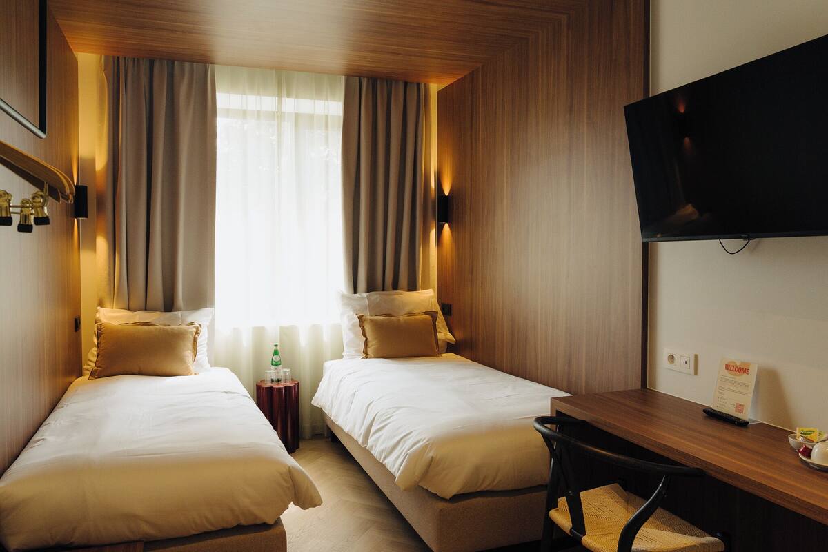 A hotel room with two beds, a desk, a chair, a TV, and a window with curtains.