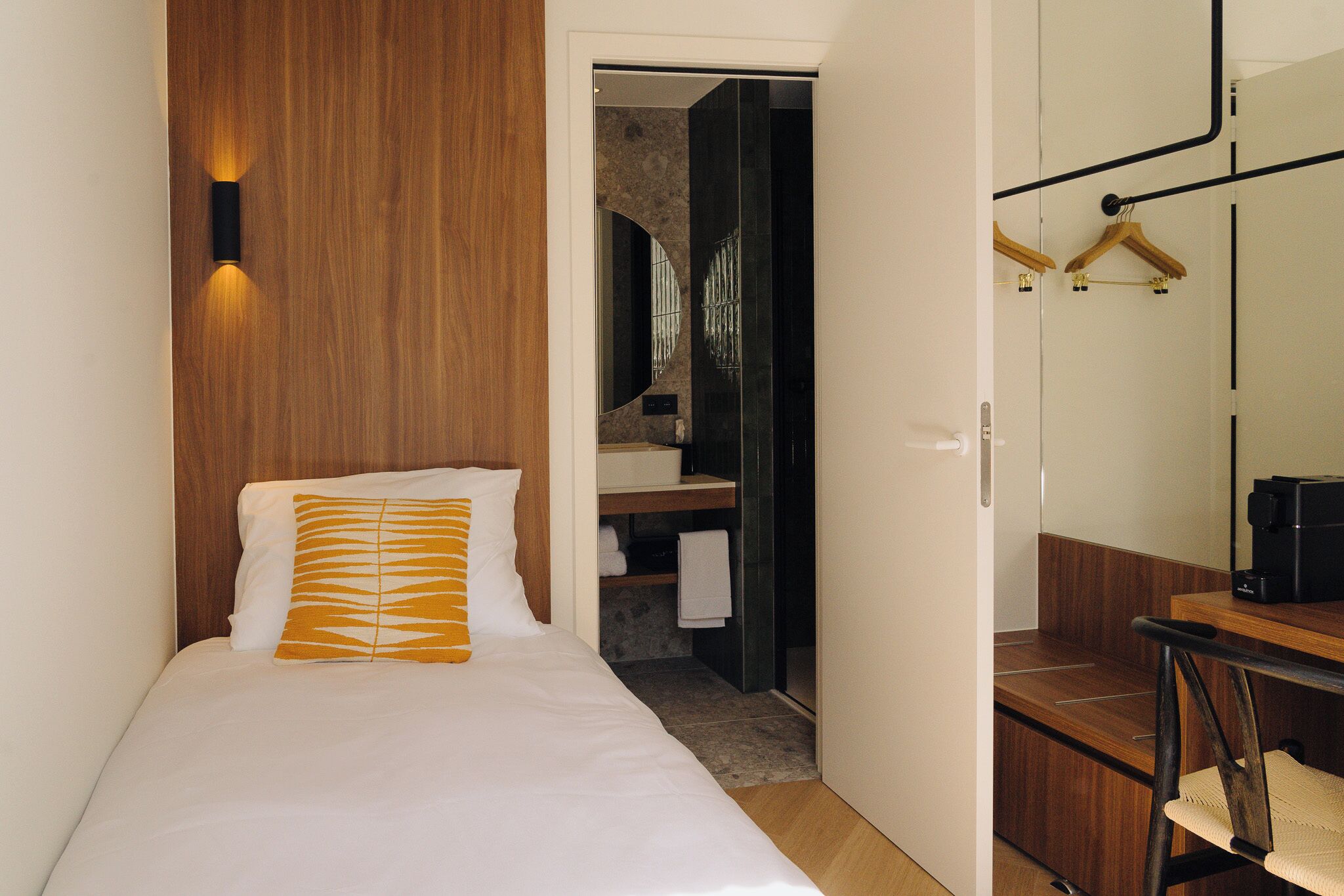 A hotel room with a bed, a wooden headboard, a mirror, a wooden desk, and a bathroom visible through an open door.
