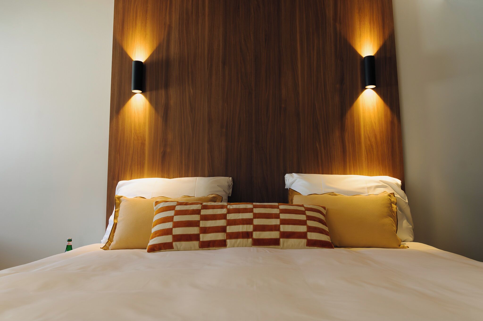 Une chambre d’hôtel avec une tête de lit en bois, deux lampes de chevet, un plaid à carreaux et une bouteille de champagne posée sur la table de chevet.