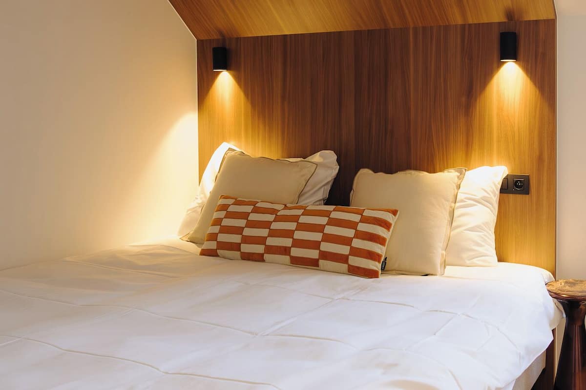 A bedroom with a wooden sloped ceiling, a bed with white bedding, two wall-mounted lights, and a small bedside table.