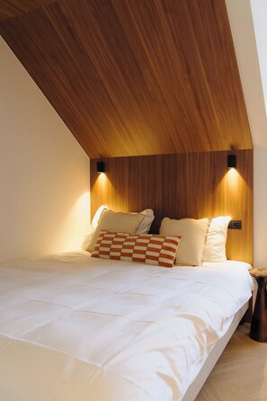 A bedroom with a wooden sloped ceiling, a bed with white bedding, two wall-mounted lights, and a small bedside table. - Door9 (Hasselt)