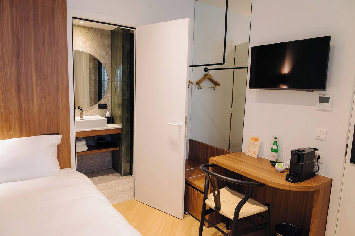 A hotel room with a bed, a desk with a chair, a TV, a mirror, and a bathroom with a sink and shower.