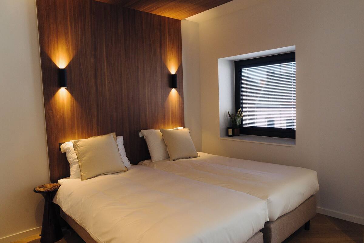 A bedroom with a large bed, two bedside tables, a wooden headboard, and a window with blinds.