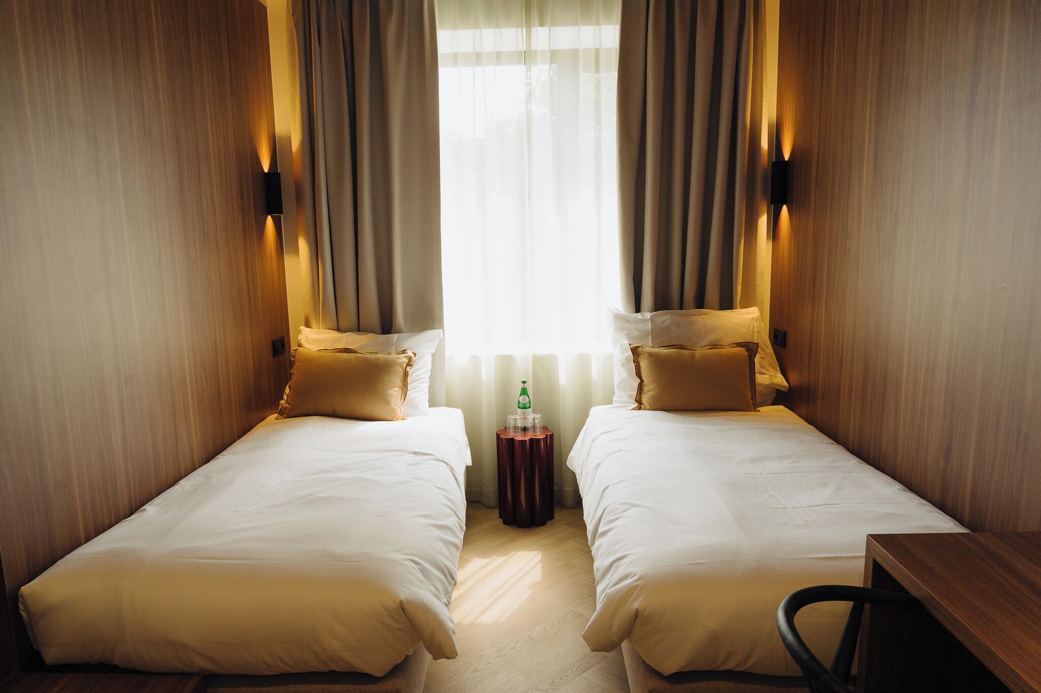 Une chambre d’hôtel avec deux lits simples, une petite table ronde et une chaise. Il y a une fenêtre avec des rideaux et un luminaire fixé au mur.