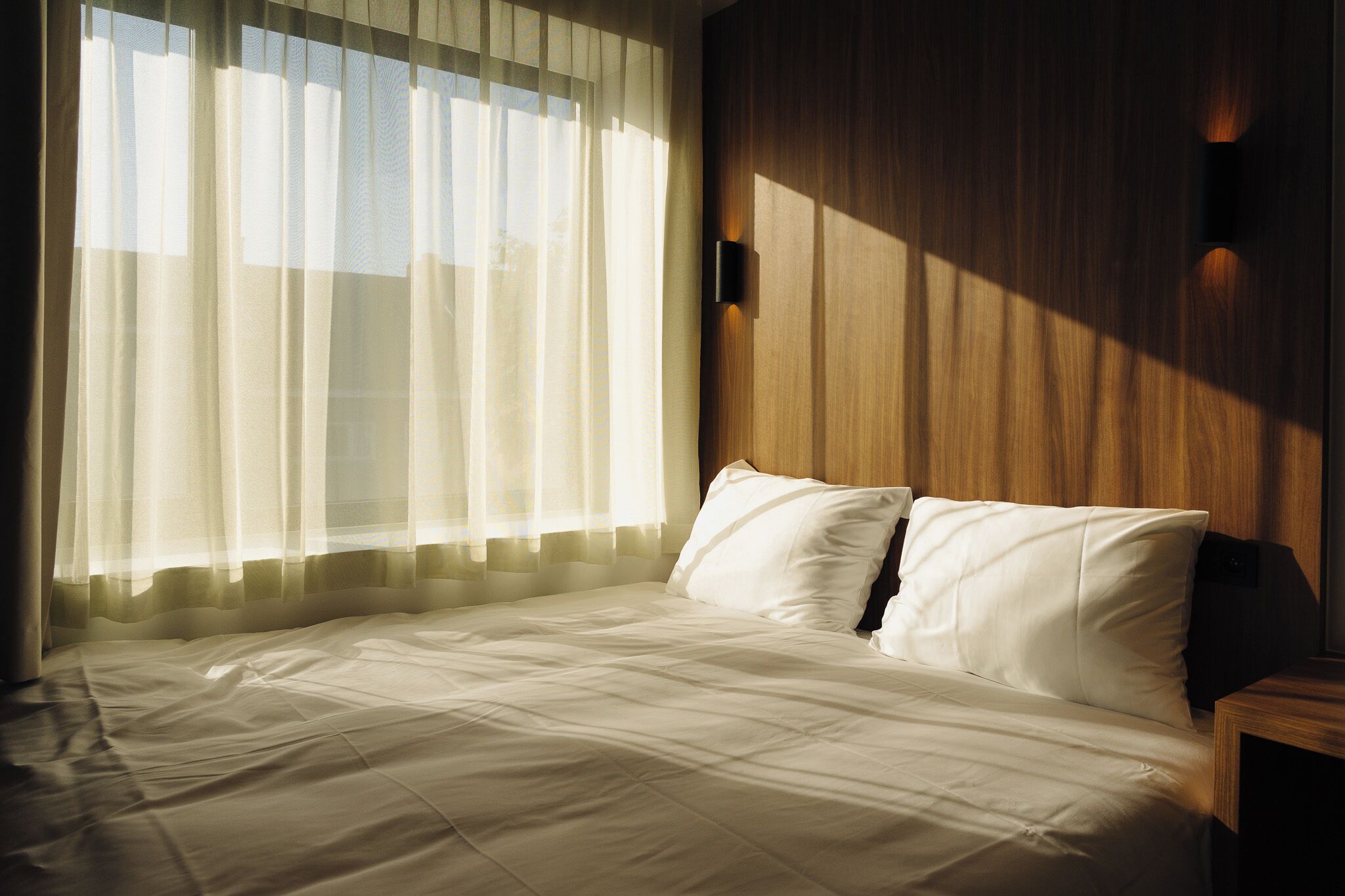 A neatly made bed with white linens, a wooden headboard, and a wall-mounted light fixture.