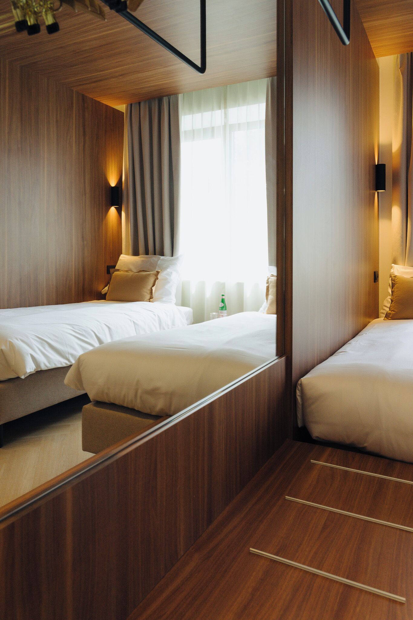 A hotel room with two beds, wooden paneling, a window with curtains, and a nightstand with a bottle.