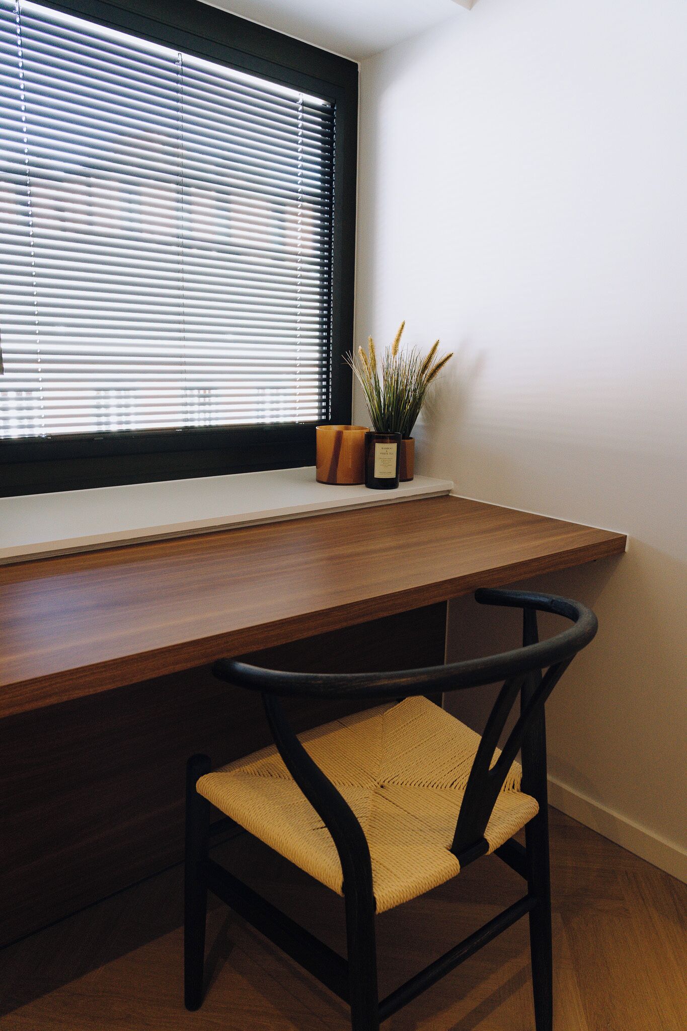Een houten bureau met een stoel, een raam met zonwering en een bloempot op de vensterbank.