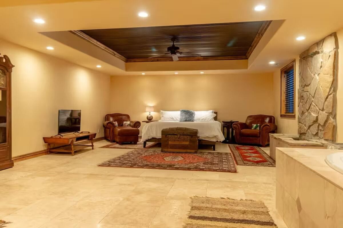 A spacious bedroom with a large bed, two leather chairs, a wooden bench, a stone accent wall, and a stone fireplace.