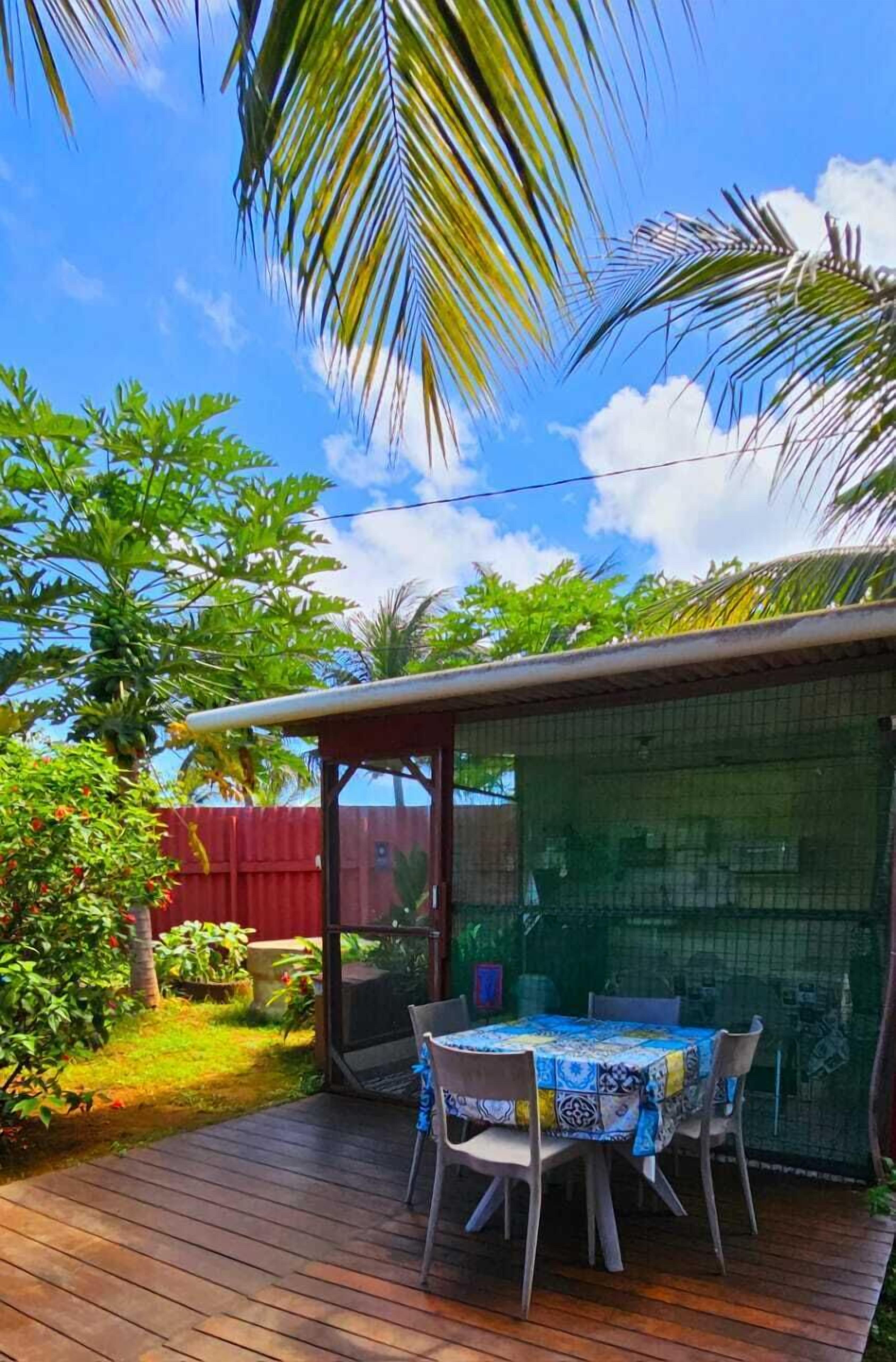Une terrasse en bois avec une table et des chaises, entourée d’une végétation luxuriante et d’une clôture rouge.