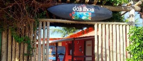 A wooden gate with a sign reading "das ilhas" leading to a building with a red roof and a visible pool area.