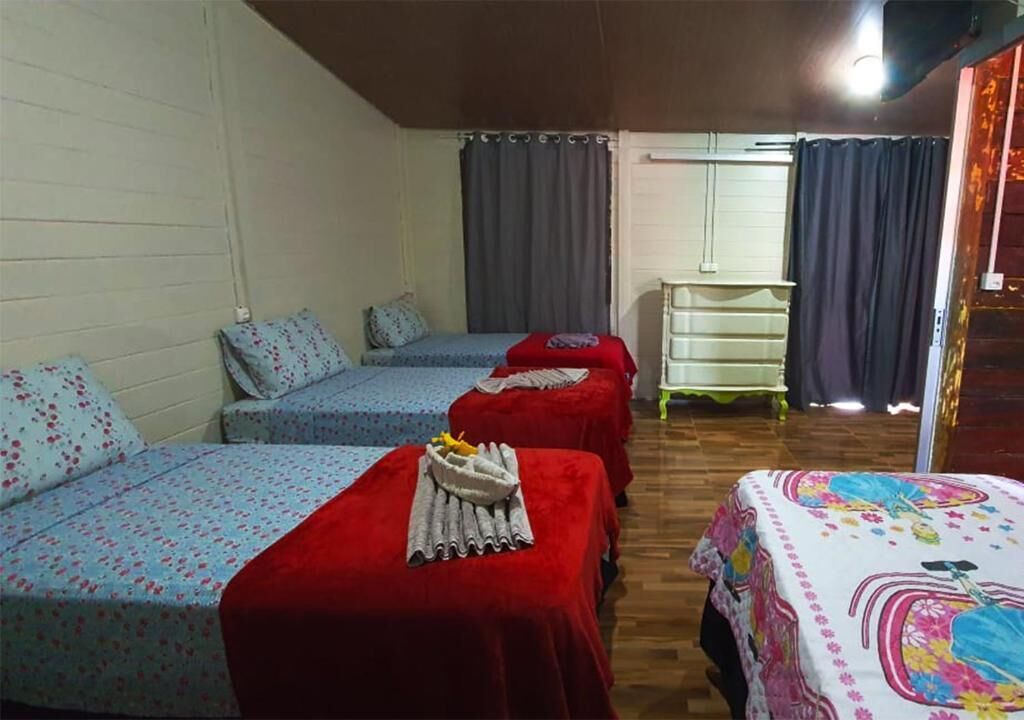 Habitación con tres camas, cada una con una colcha de diferente color. En el centro, una mesita con una toalla y un frutero.