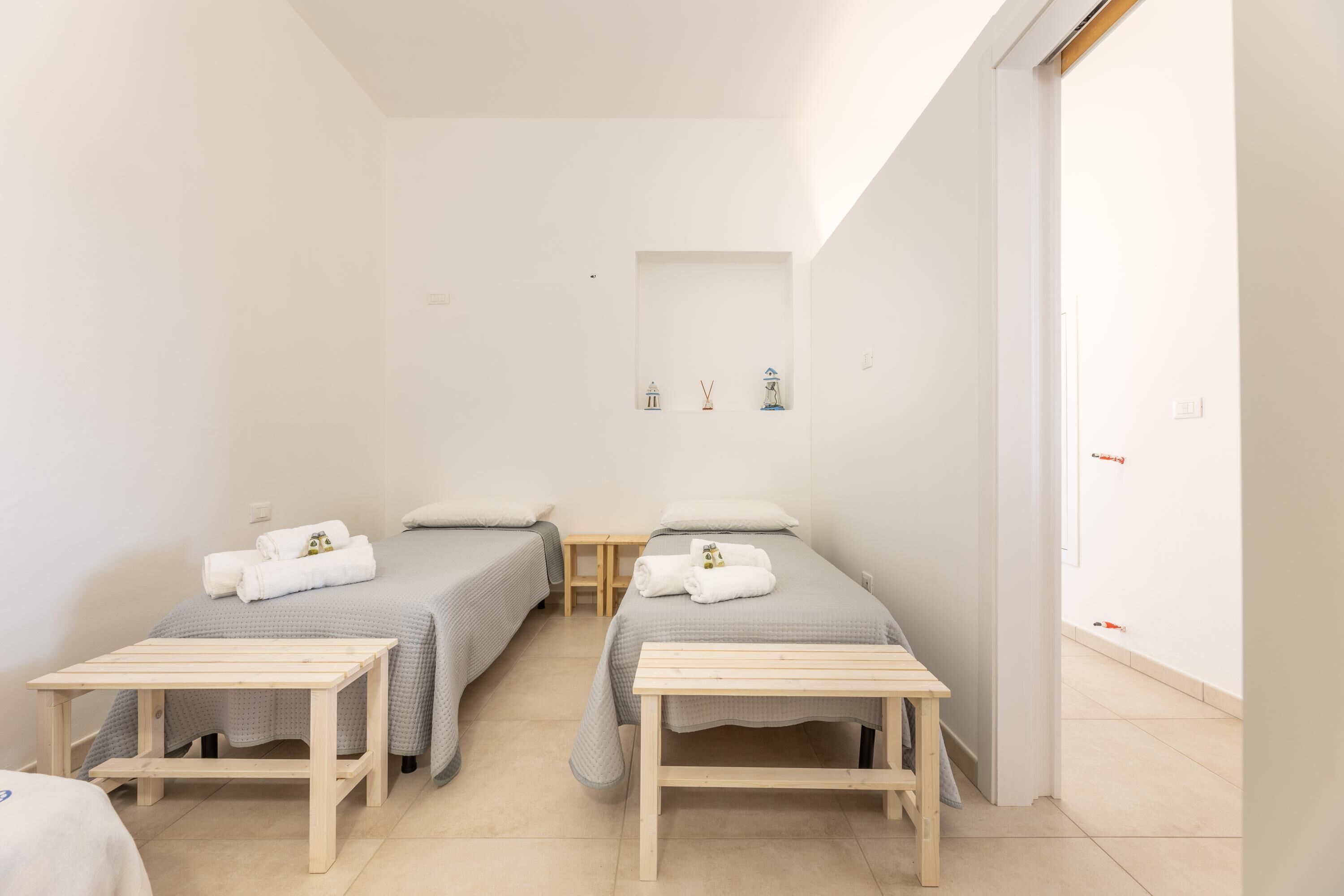 A small, clean room with two single beds, a wooden table, and a small shelf with decorative items.