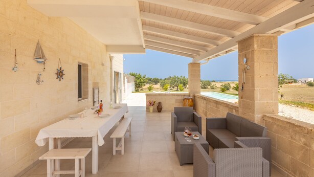 A covered outdoor seating area with a table and benches, overlooking a pool and a scenic landscape. - Villa Serenella - Carovigno (Carovigno)