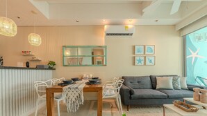A modern living room with a grey sofa, white chairs, a wooden dining table, and a kitchen area with a striped wall and hanging lights. - Edificio Orange Castillogrande (Cartagena)