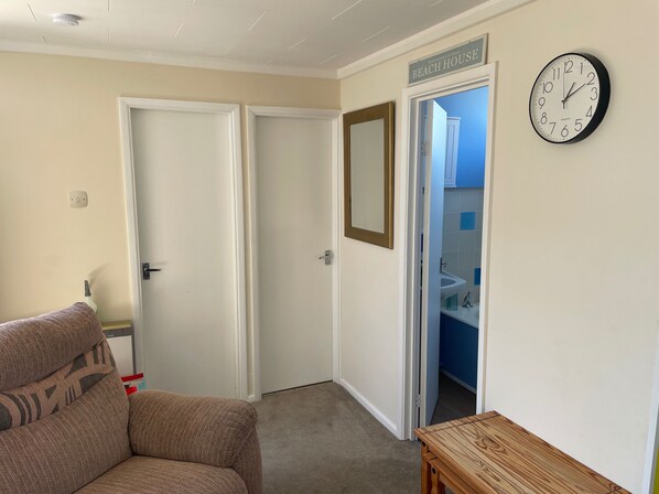 A room with a beige carpet, a brown sofa, a wooden table, a clock, and a door leading to a bathroom with a sink and blue tiles. - 2 Bed Lodge Sleeps 4- Pets, Parking, 2min to Beach (Skegness)
