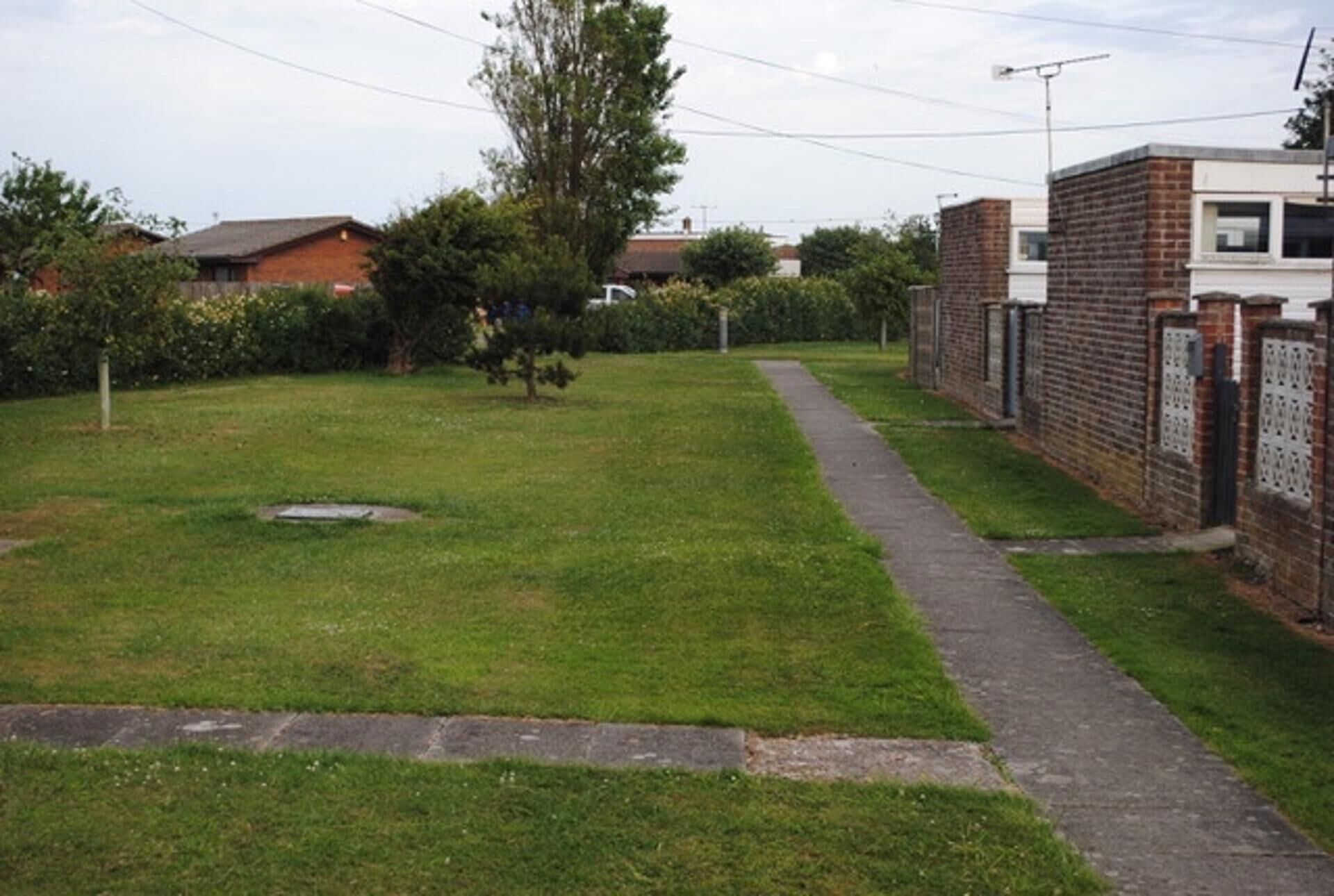 A grassy area with a paved path, a brick wall, and a tree.