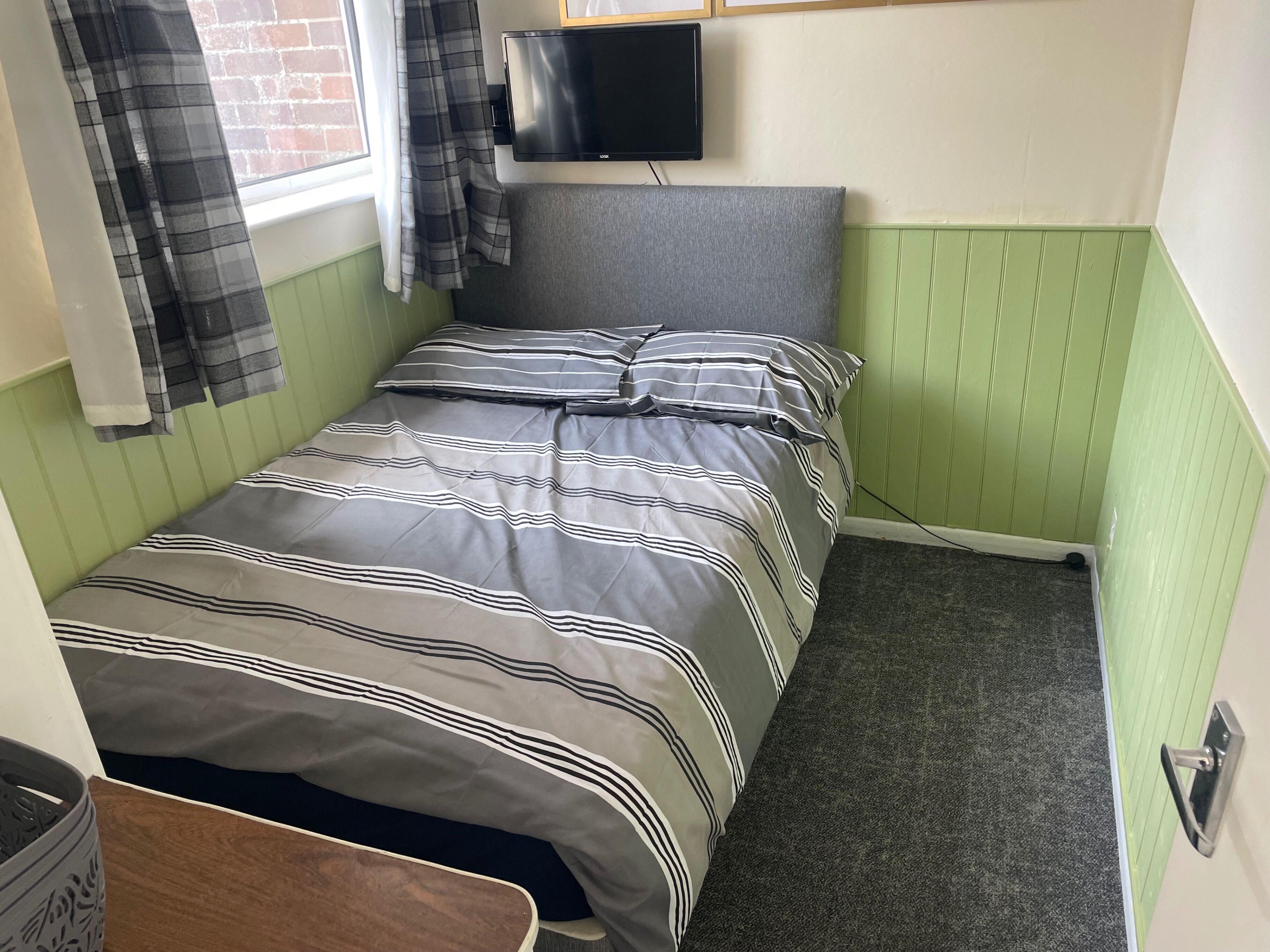A small, neatly arranged bedroom with a bed, a television mounted on the wall, and a window with curtains.