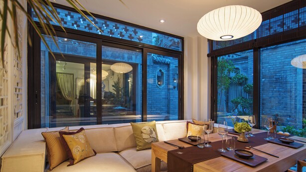 A modern dining area with a wooden table set for a meal, a sofa with cushions, and large windows offering views of the outside.