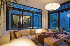 A modern dining area with a wooden table set for a meal, a sofa with cushions, and large windows offering views of the outside. - Beijing 161 Lezai Nanluo Hotel (Beijing)