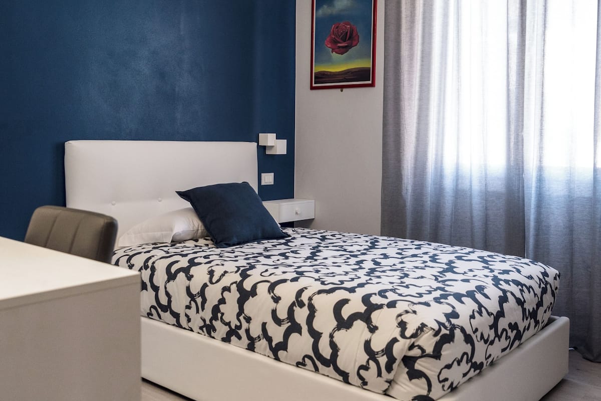 Una camera da letto moderna con un letto bianco, una parete azzurra, un quadro incorniciato e una finestra con tende.