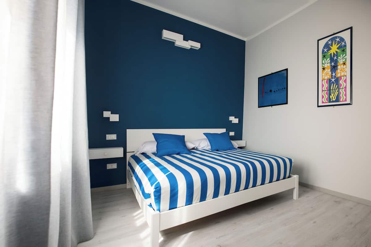 Una camera da letto con un letto bianco, biancheria a righe blu e bianche, una parete azzurra a contrasto e un'opera d'arte colorata appesa al muro.
