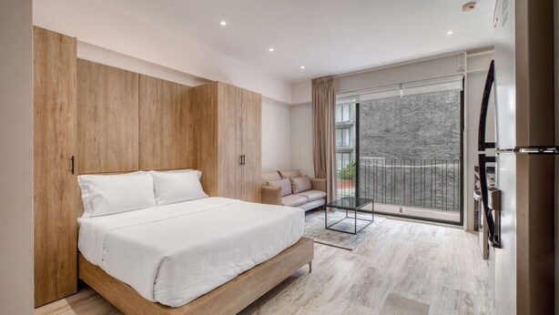 A modern bedroom with a wooden bed frame, a sofa, and a balcony with a view. - Myla - Loft 09 - Chapultepec LowF (Mexico City)