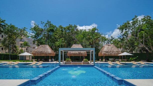 Piscina con sillones de playa y sombrillas de paja, rodeada de árboles y edificios.