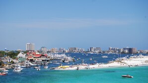 Un paisaje urbano costero con un puerto deportivo lleno de barcos, una playa arenosa y diversos edificios, incluyendo hoteles y viviendas.