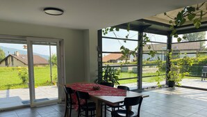 A dining area with a table set for four, surrounded by chairs, and a view of a garden and buildings through large windows. - Bell Air near Neuchatel (Val-de-Ruz)