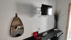 Une pièce avec un téléviseur à écran plat fixé au mur, un miroir, une petite étagère sur laquelle se trouve une boîte rouge et un vase contenant de longues tiges.