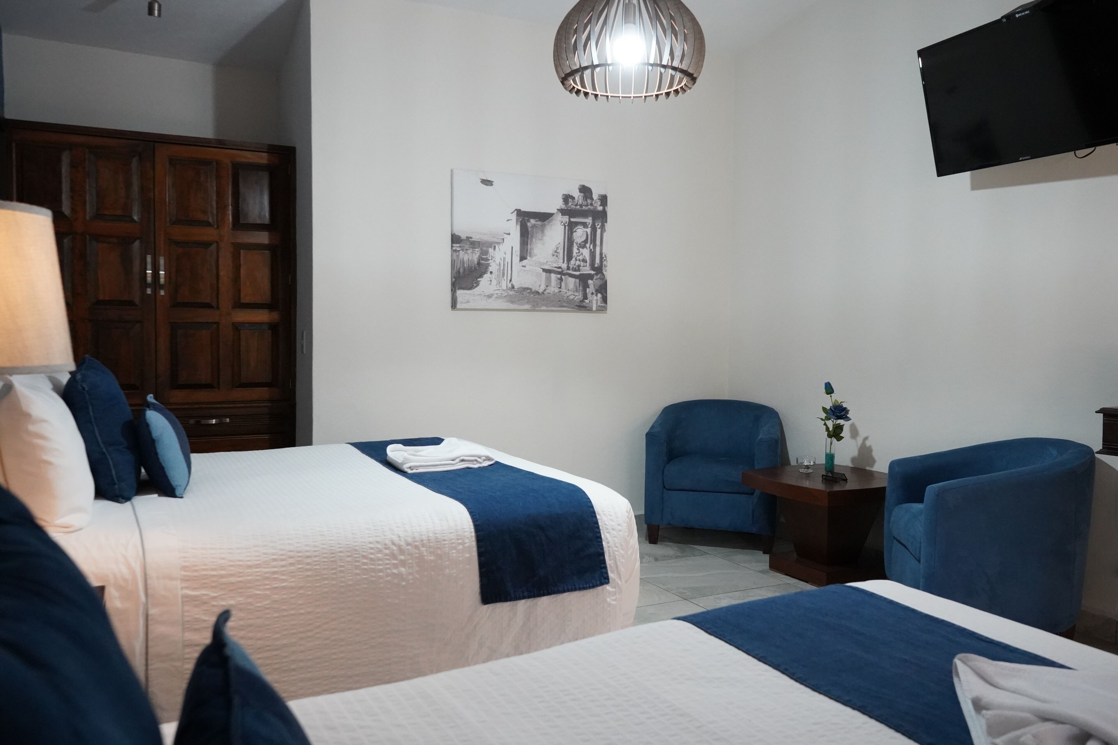 Habitación de hotel con dos camas, un armario de madera, un sillón azul, una mesita con un jarrón y un televisor montado en la pared.