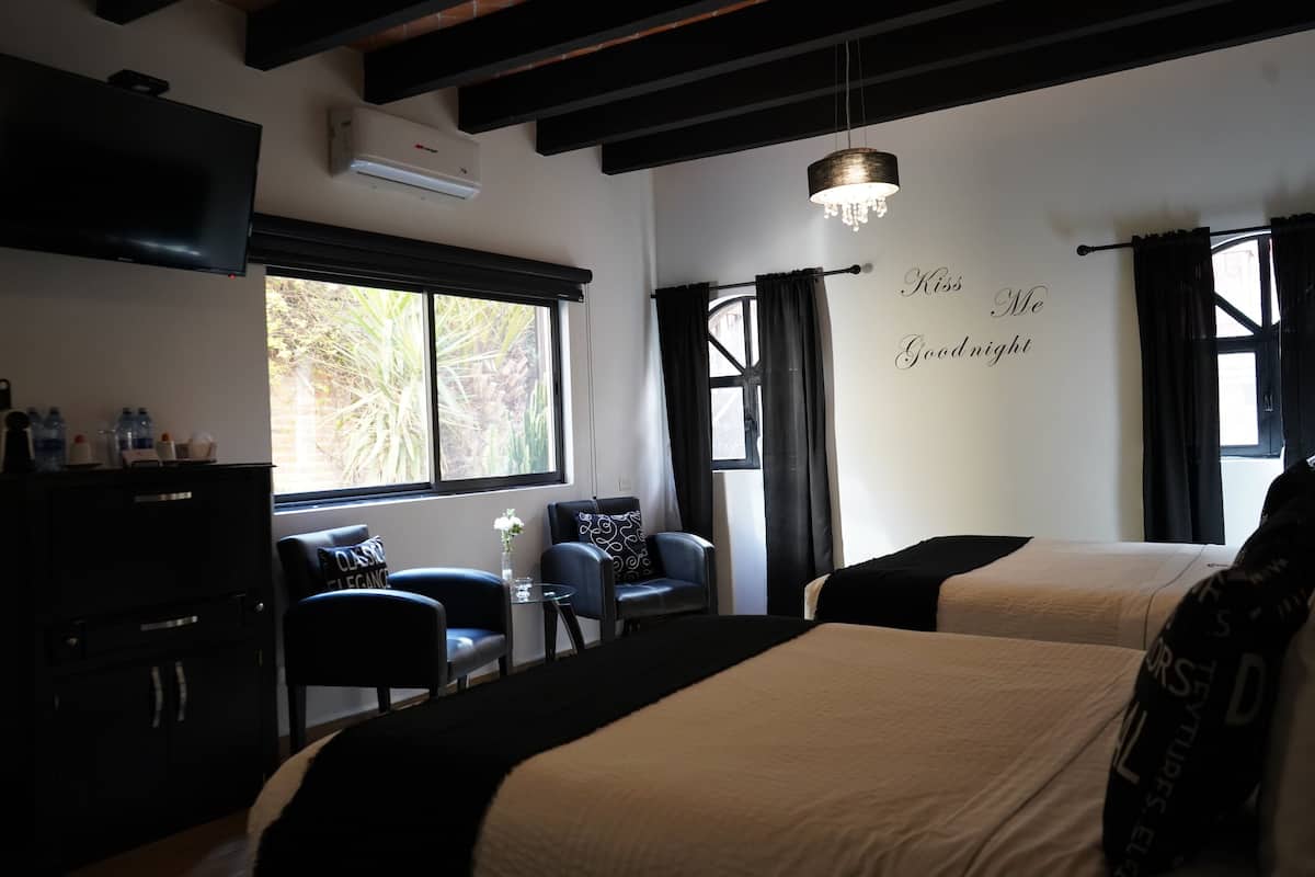 Quarto de hotel com duas camas, televisão, escrivaninha com cadeiras e janela com cortinas.