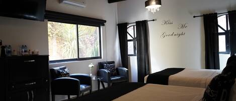 A hotel room with two beds, a TV, a desk with chairs, and a window with curtains.