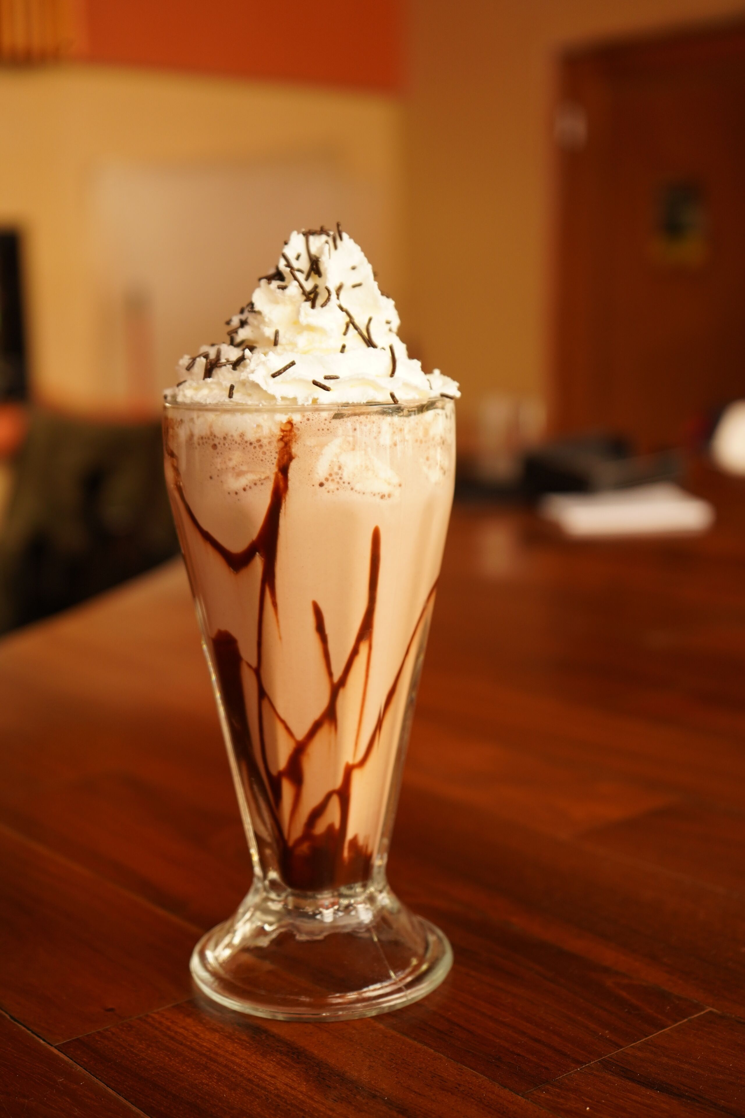 A tall glass of chocolate milkshake with whipped cream and chocolate syrup on a wooden table.
