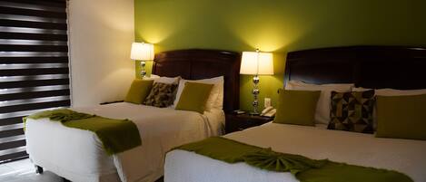 Two beds with white linens and green throw blankets, each with a bedside table and lamp.