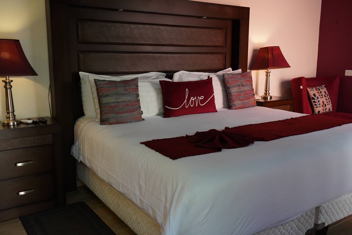 Cama bem arrumada com lençóis brancos e manta vermelha, acompanhada por travesseiros decorativos, incluindo um com a palavra 'love'.