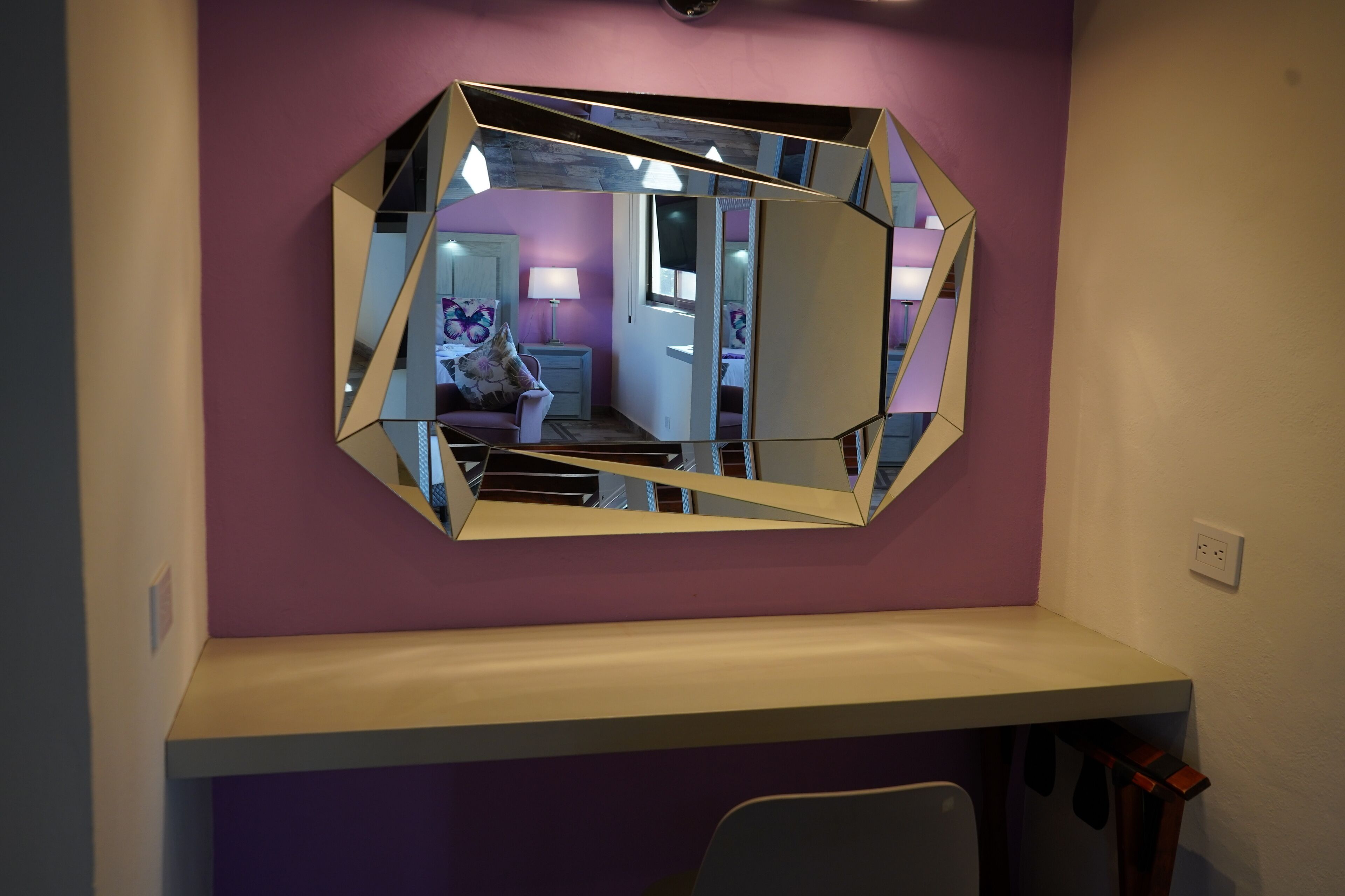 A room with a large, geometric mirror on the wall, a purple accent wall, and a white shelf below.