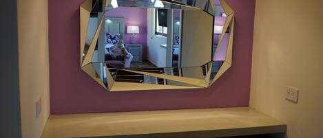 A room with a large, geometric mirror on the wall, a purple accent wall, and a white shelf below.