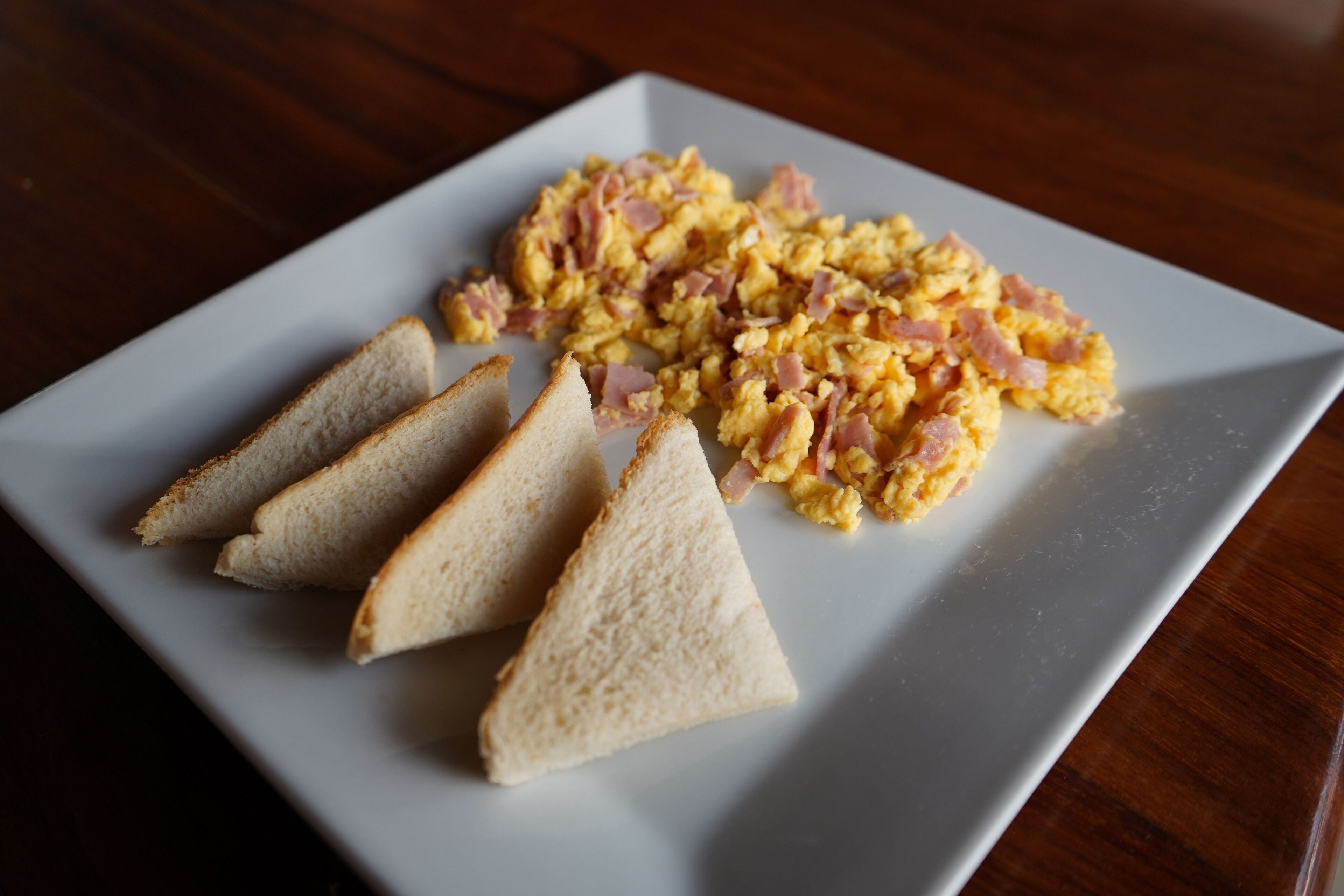 A white plate with scrambled eggs and ham, accompanied by three slices of toast on a wooden table.