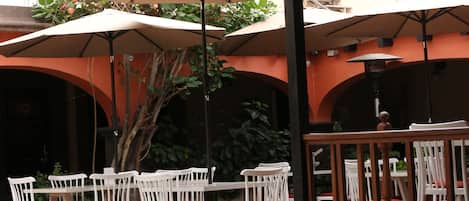 An outdoor patio area with white chairs and tables, covered by umbrellas, and surrounded by orange walls.