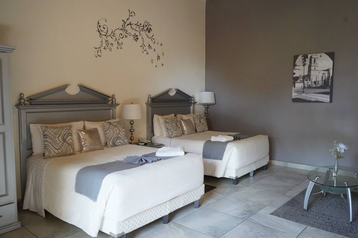 Quarto de hotel com duas camas, uma mesa pequena e um objeto decorativo na parede.