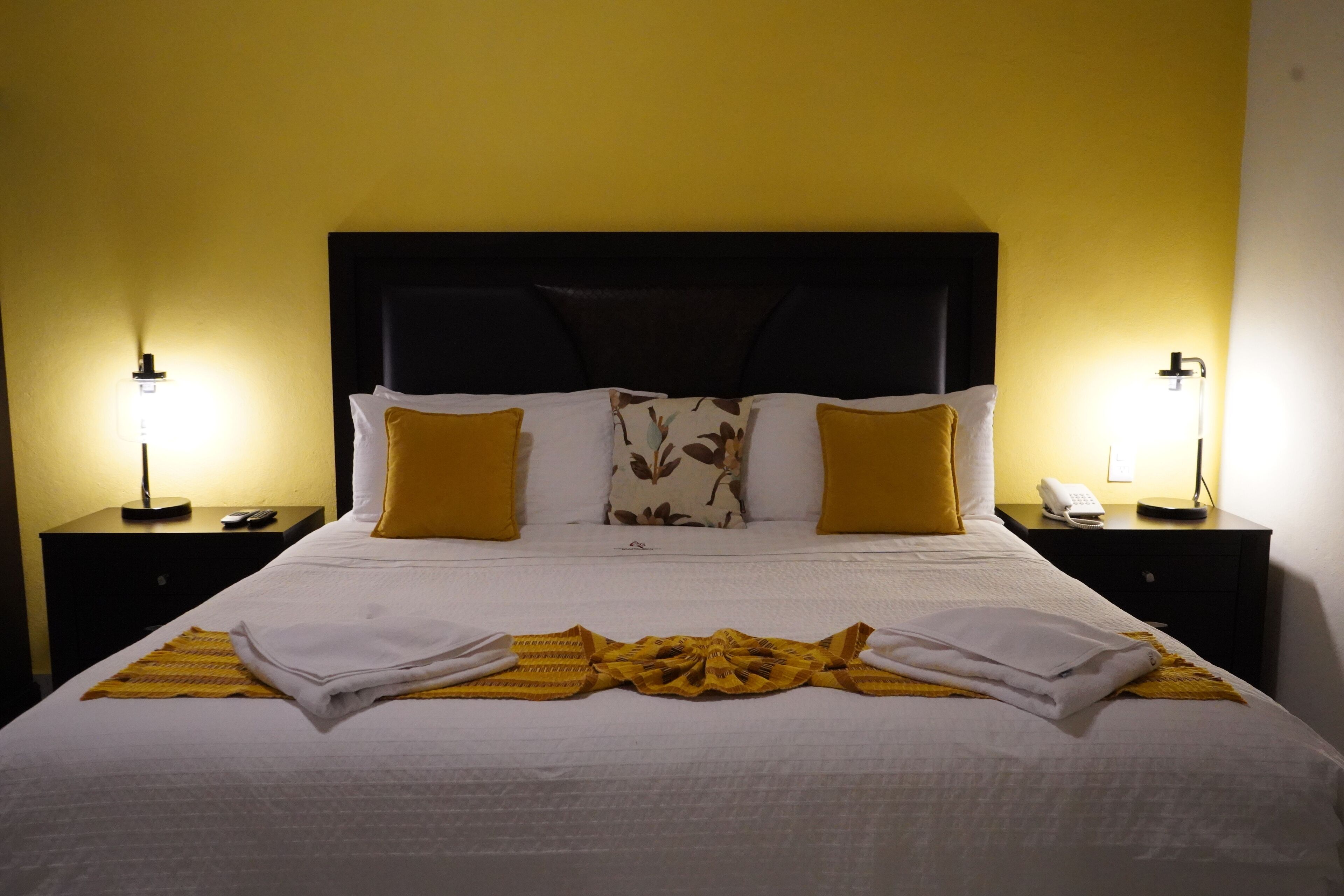 Una cama bien tendida con sábanas blancas y una manta amarilla, flanqueada por dos mesitas de noche con lámparas y un teléfono.