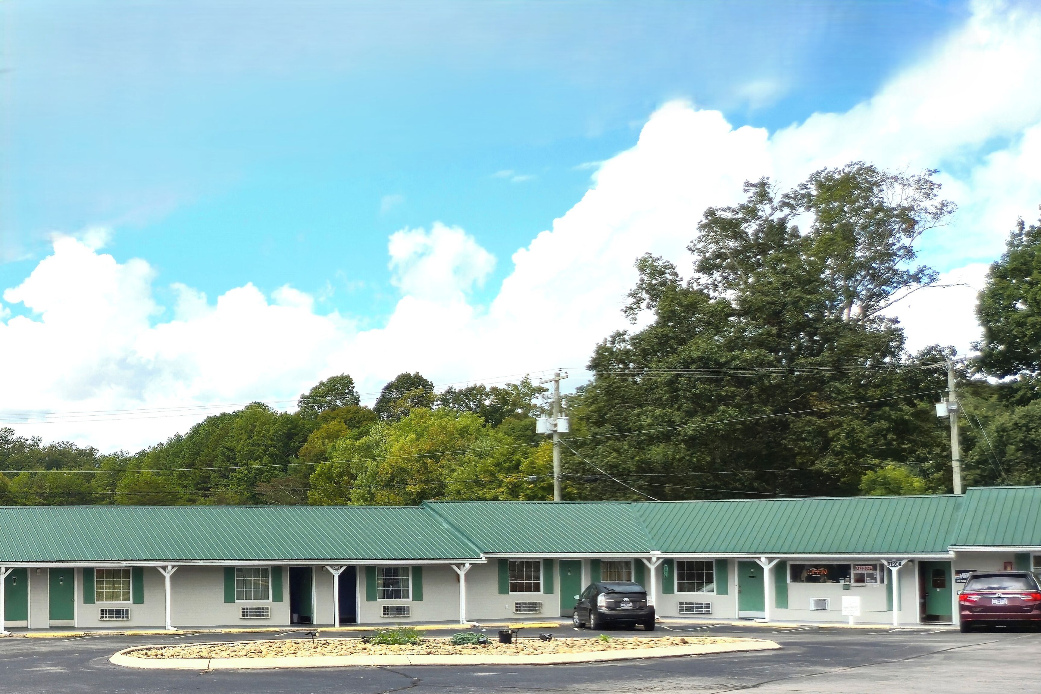 A motel with a green roof and white exterior, featuring a parking lot and a few parked cars.