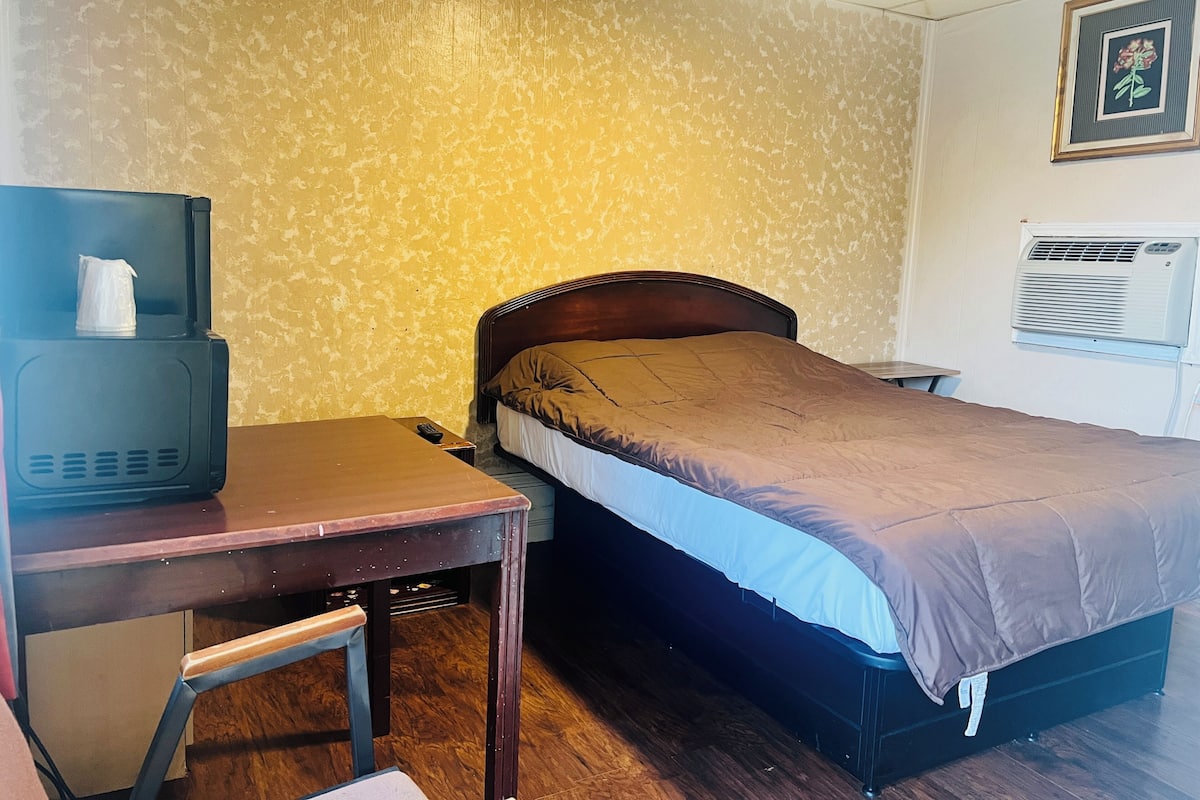 Hotellihuone, jossa on sänky, työpöytä, jossa on televisio, tuoli ja ilmastointilaite.