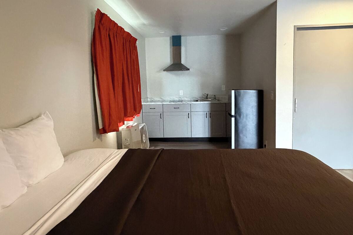 A hotel room with a bed, a small kitchenette with a refrigerator, and a window with red curtains.