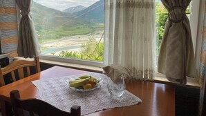 A table with a bowl of fruit and a glass vase, set against a scenic view of mountains and a river through a window with curtains. - Vila across Tepelena (Tepelenë)