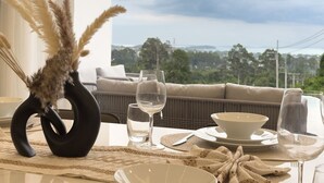 Une table à manger est dressée avec de l’assiette blanche, des bols et des verres à vin, et un vase noir contient des plumes d’herbe des pampas. Un canapé et une vue sur la verdure se trouvent à l’arrière-plan.