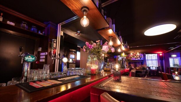 Una zona de bar con mostrador de madera, taburetes y diferentes tipos de vasos.