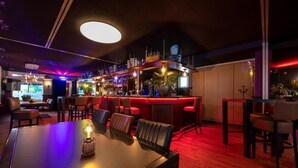 Zona de bar con una mesa larga y varios taburetes, una barra con un cantinero, y un área de estar con sillas y mesas.